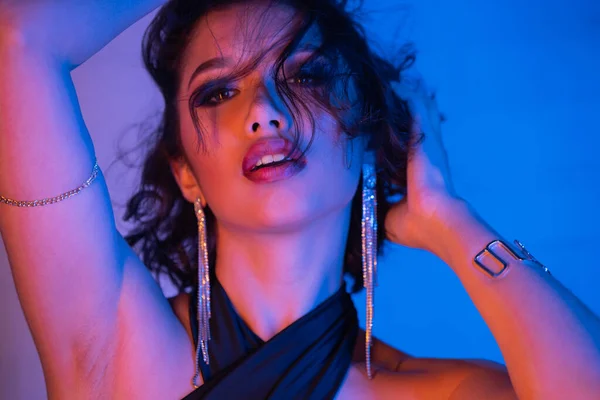 Confiante jovem mulher asiática na roupa da moda tocando o cabelo e de pé na luz do néon no clube noturno — Fotografia de Stock