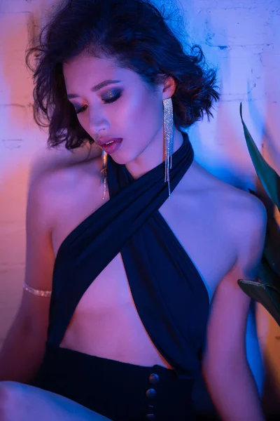 Atractiva joven mujer asiática en vestido sexy sentado cerca de la planta en luz de neón en el club nocturno - foto de stock