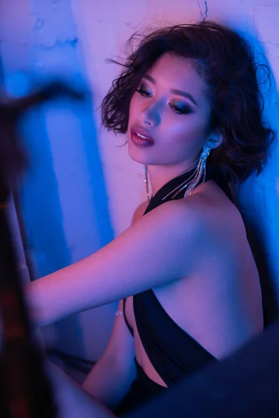 Уверенная и стильная молодая азиатка в платье, стоящая в неоновом свете в ночном клубе — стоковое фото
