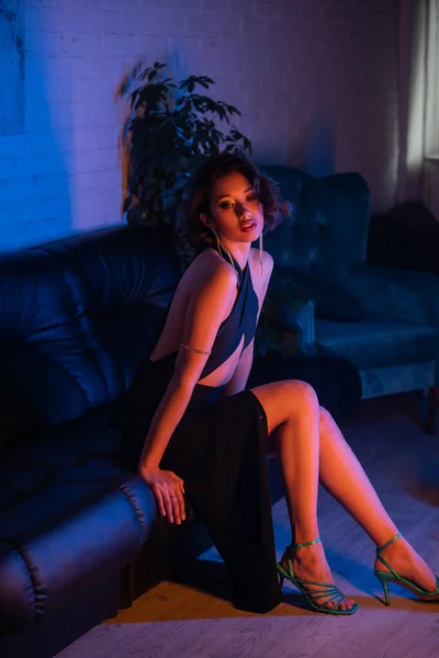 Elegante y hermosa mujer asiática en vestido sentado en el sofá en la iluminación de neón en el club nocturno - foto de stock
