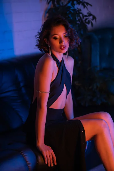 Moda asiático mujer en pendientes y vestido sentado en sofá en neón luz en noche club - foto de stock