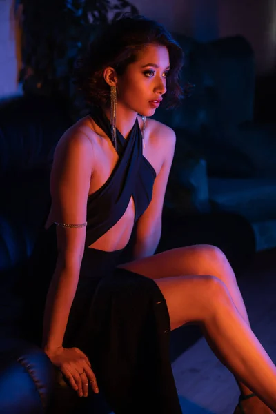 Уверенная молодая азиатка в платье и серьгах сидит на диване в ночном клубе с неоновым освещением — стоковое фото