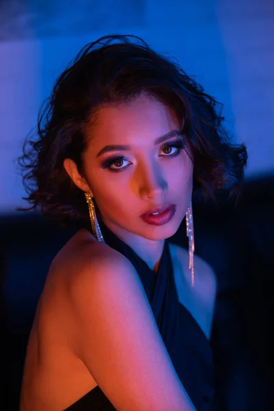 Na moda asiático mulher no vestido olhando para câmera no sofá no azul e rosa néon luz no noite clube — Fotografia de Stock