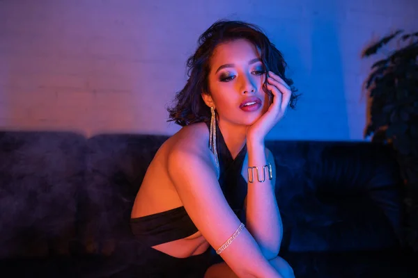 Schöne asiatische Frau im Abendkleid berührt Gesicht und sitzt auf Couch im Neon-Licht in Nachtclub — Stockfoto