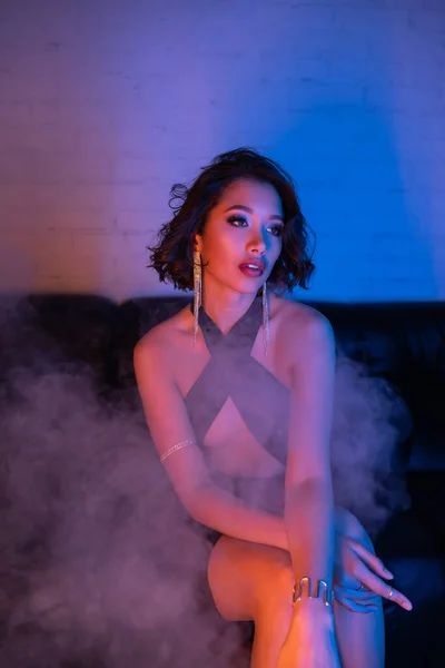 Elegant young asian woman in evening wear sitting in smoke and neon light on couch in night club — Stock Photo