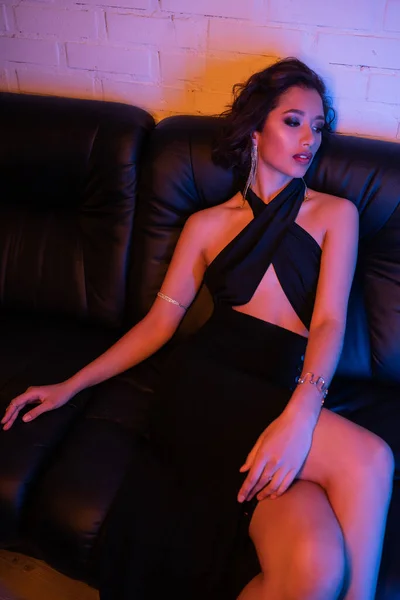 Vue grand angle de la femme asiatique à la mode en robe assise sur le canapé dans l'éclairage au néon dans la boîte de nuit — Photo de stock