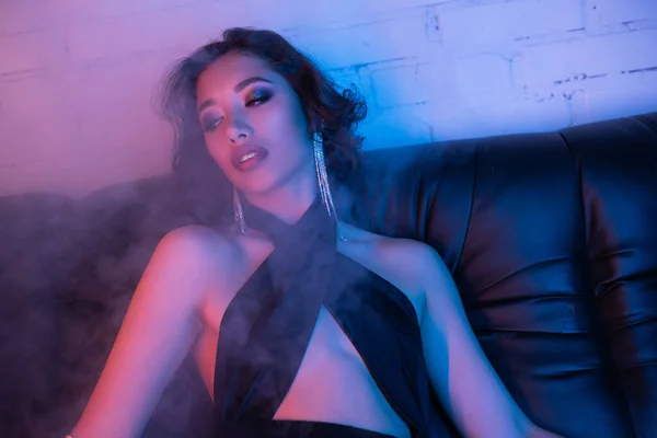 Sexy donna asiatica in abito da sera seduta in fumo e luce al neon colorata sul divano nel night club — Foto stock