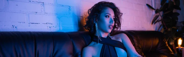 Sexy young asian woman in evening outfit sitting on couch in neon lighting in night club, banner — Stock Photo