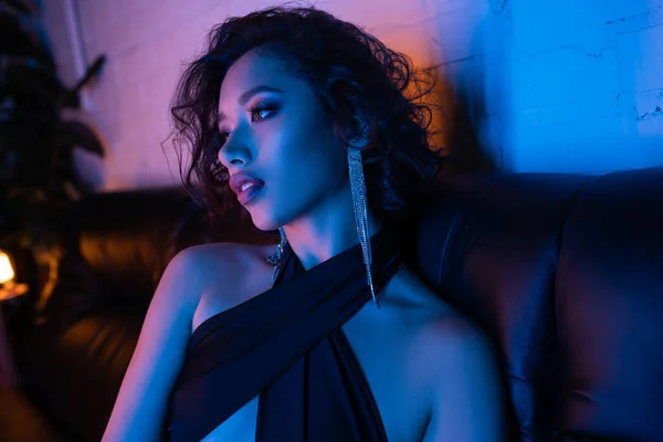 Ritratto di donna asiatica sicura di sé in abito da sera seduta sul divano in luce al neon nel night club — Foto stock