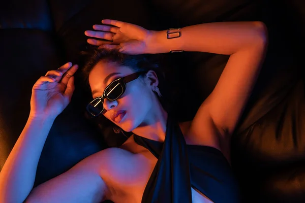 Вид сверху на молодую женщину в солнцезащитных очках и платье, лежащее на диване при неоновом свете в ночном клубе — стоковое фото