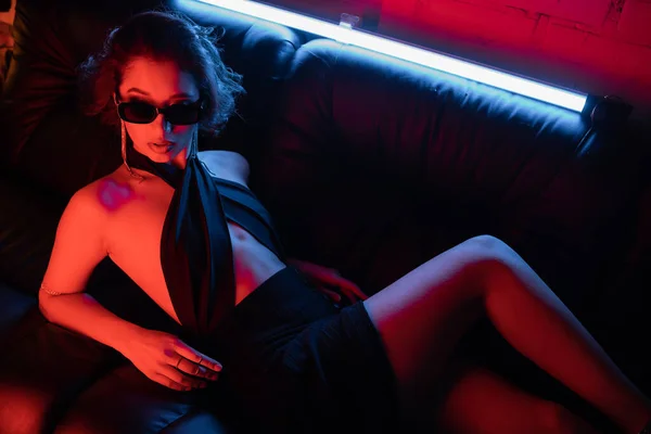 Вид стильной женщины в солнцезащитных очках, лежащей в неоновом свете на диване в ночном клубе — стоковое фото