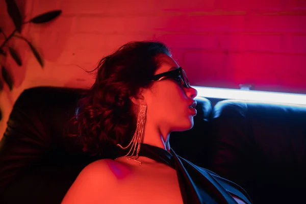 Vista lateral de sexy mujer asiática en gafas de sol y pendiente posando en sofá en luz de neón en club nocturno - foto de stock