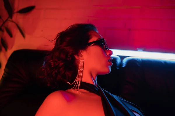 Seitenansicht von sexy asiatische Frau mit Sonnenbrille und Ohrring posiert auf Couch im Neonlicht in Nachtclub — Stockfoto