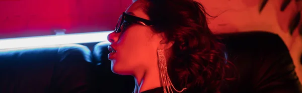 Vista laterale della donna asiatica alla moda con gli occhiali da sole seduta sul divano nella luce al neon nel night club, banner — Foto stock