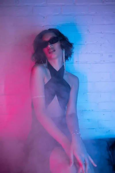 Sexy mujer asiática en gafas de sol y vestido sentado en el sofá en el humo y luz de neón en el club nocturno - foto de stock