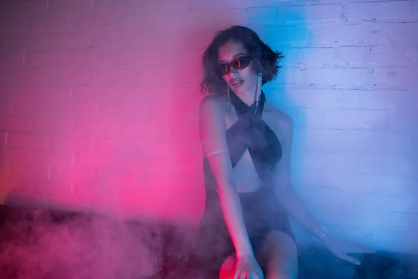 Joven mujer asiática en gafas de sol sentado en el sofá durante la fiesta en el humo y la luz de neón en el club nocturno - foto de stock