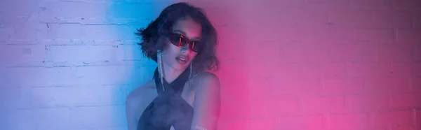 Молодая азиатка в солнечных очках позирует в дыму и неоновом освещении в ночном клубе, баннер — стоковое фото