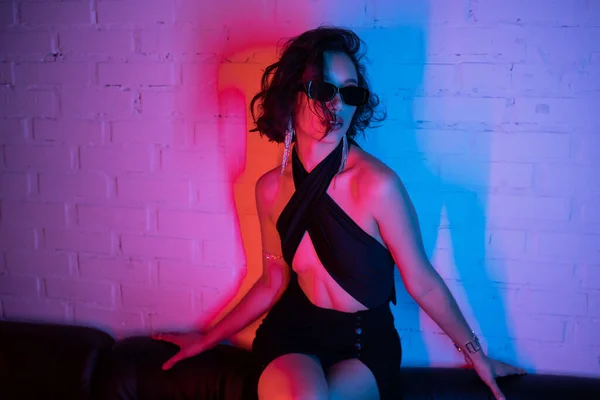 Сексуальная азиатка в солнечных очках, позирующая в розовом и синем неоновом свете на диване в ночном клубе — стоковое фото