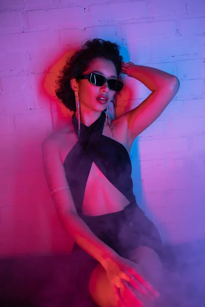 Mujer asiática con estilo en gafas de sol tocando el pelo y sentado en el humo y la luz de neón en el club nocturno — Stock Photo