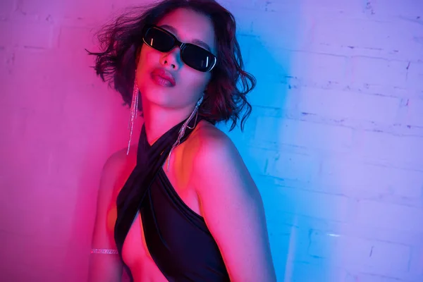 Sexy young asian woman in sunglasses posing in vibrant neon light and smoke in night club — Stock Photo