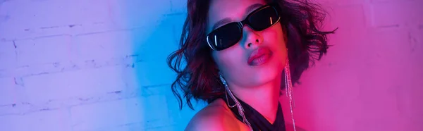 Модная молодая женщина в солнцезащитных очках позирует при ярком неоновом освещении в ночном клубе, баннер — Stock Photo