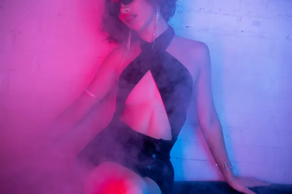 Cropped view of sexy woman in sunglasses sitting on couch in smoke and neon light in night club — Stock Photo