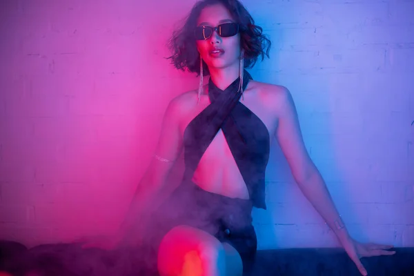 Mujer asiática de moda en vestido sexy y gafas de sol posando en humo y luz de neón en club nocturno - foto de stock