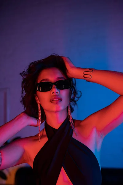 Sexy young asian woman in sunglasses touching hair while standing in vivid neon light in night club — Stock Photo