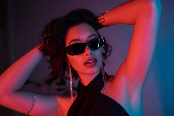 Sexy asian woman in sunglasses and bracelets touching head in neon light in night club — Stock Photo