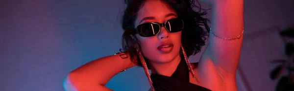 Trendy asian woman in sunglasses and bracelets posing in colorful neon light in night club, banner — Stock Photo