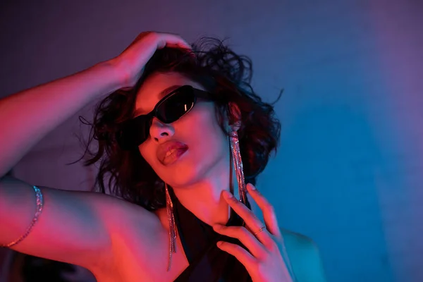 Giovane donna asiatica in occhiali da sole toccare la testa mentre in piedi in luce al neon colorato in night club — Foto stock