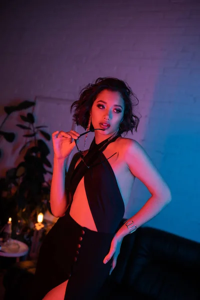 Сексуальная азиатка в платье, держащая солнцезащитные очки и глядя в ночной клуб с неоновым светом — стоковое фото