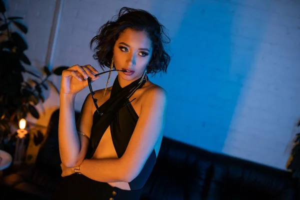 Mujer asiática segura y sexy sosteniendo gafas de sol mientras está de pie en el club nocturno con luz de neón - foto de stock