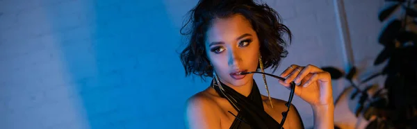 Sexy junge asiatische Frau mit Make-up hält Sonnenbrille in Nachtclub mit Neonlicht, Banner — Stockfoto