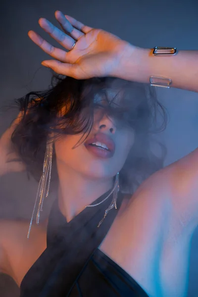 Elegante mujer asiática en vestido y pendientes bailando en humo y luz de neón en club nocturno - foto de stock