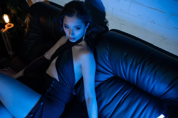 Vue grand angle de la jeune femme asiatique élégant assis sur le canapé dans la lumière néon bleu dans la boîte de nuit — Photo de stock