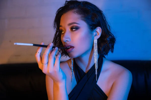 Moda jovem mulher asiática com maquiagem segurando cigarro no bocal em néon luz no clube noturno — Fotografia de Stock