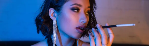 Trendy asiatische Frau mit Visage hält Zigarette im Mundstück in Nachtclub mit Neonlicht, Banner — Stockfoto