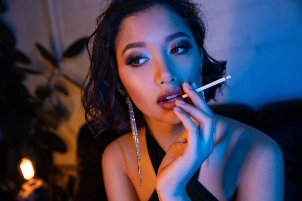 Atractiva joven asiática mujer en vestido de fumar cigarrillo en club nocturno con luz de neón - foto de stock