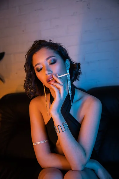 Elegante y elegante mujer asiática fumando cigarrillo mientras pasa tiempo en el club nocturno con luz de neón - foto de stock