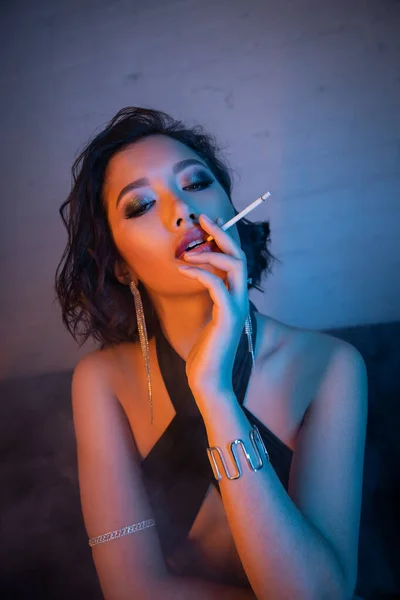 Retrato de sexy mujer asiática en vestido y pulseras fumar cigarrillo en luz de neón en club nocturno - foto de stock