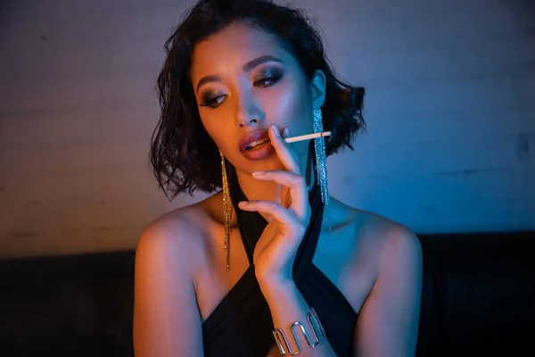 Sexy young asian woman in dress smoking cigarette and spending time in night club with neon light — Stock Photo