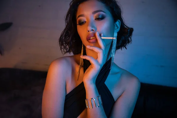 Selbstbewusste und sexy asiatische Frau raucht Zigarette, während sie Zeit in einem Nachtclub mit Neonlicht verbringt — Stockfoto
