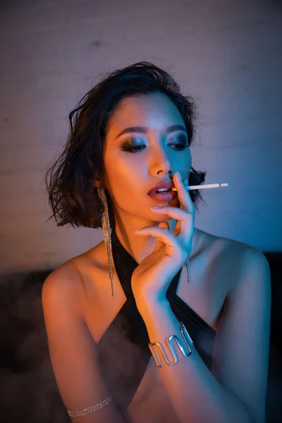 Elegante mujer asiática en vestido de fumar cigarrillos, mientras que de pie en la iluminación de neón en el club nocturno - foto de stock
