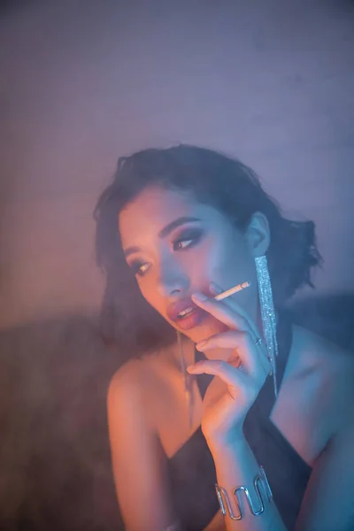 Fashionable young asian woman smoking cigarette in smoke and neon light in night club — Stock Photo