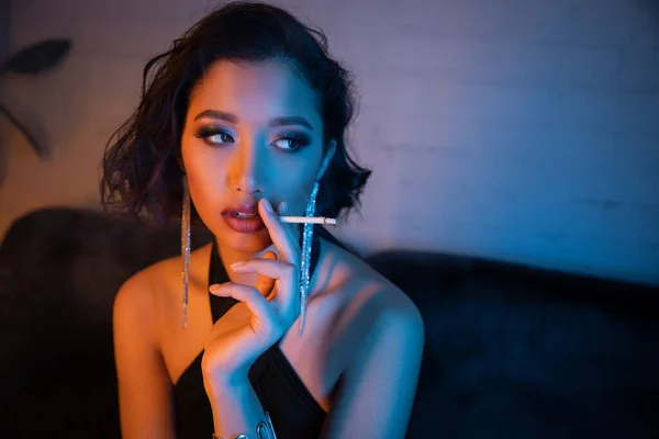Fashionable young asian woman smoking cigarette and looking away in neon lighting in night club — Stock Photo