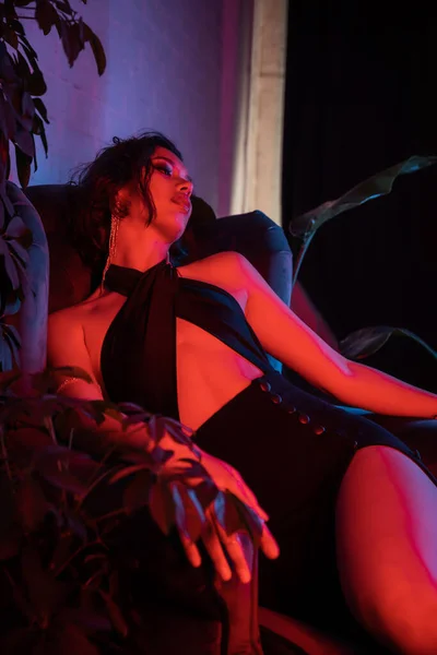 Сексуальна азіатська жінка в вечірній сукні сидить на кріслі біля рослин в нічному клубі з неоновим освітленням — стокове фото