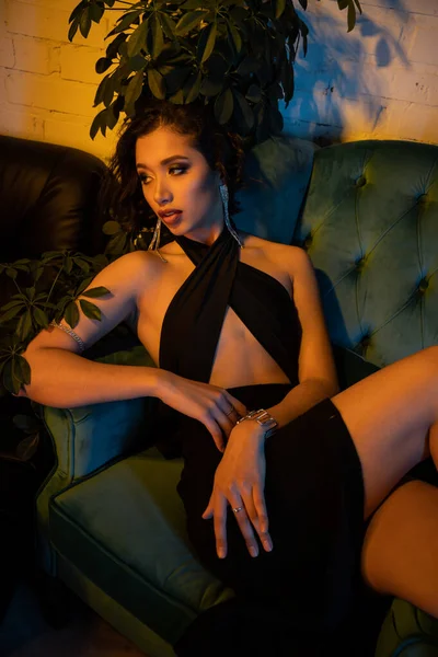 Модна азіатська жінка в вечірній сукні сидить на дивані біля рослин в нічному клубі з освітленням — стокове фото