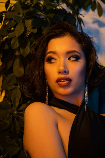 Selbstbewusste junge Asiatin mit Make-up schaut in der Nähe von Pflanzen in Nachtclub mit Beleuchtung weg — Stockfoto