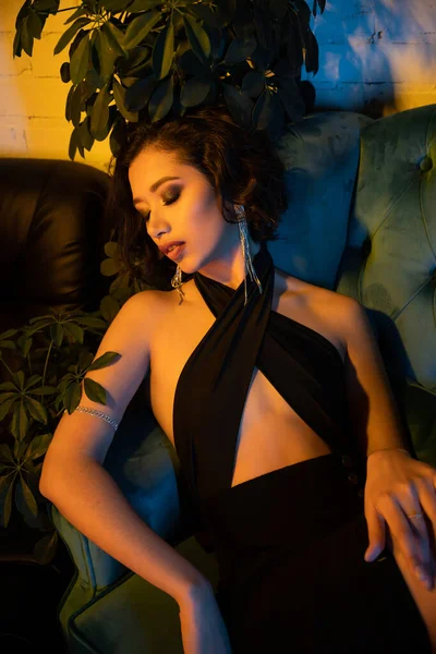 Sexy mujer asiática con maquillaje sentado en el sofá con los ojos cerrados en el club nocturno moderno con iluminación - foto de stock